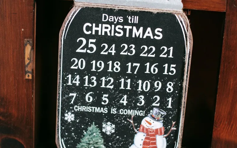 Calendrier à rebours « Days 'till Christmas » en forme de bocal, chiffres de 25 à 1, bonhomme de neige et sapin, accroché à une commode en bois