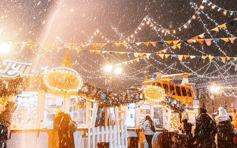 Chalets d'un marché de Noël illuminés sous une forte chute de neige, guirlandes de lumières et de fanions tendues au-dessus des passants, enseignes en cyrillique