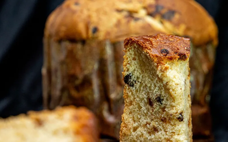 Panettone entamé, une part debout au premier plan montrant sa mie jaune parsemée de raisins secs et de fruits confits, sur fond noir