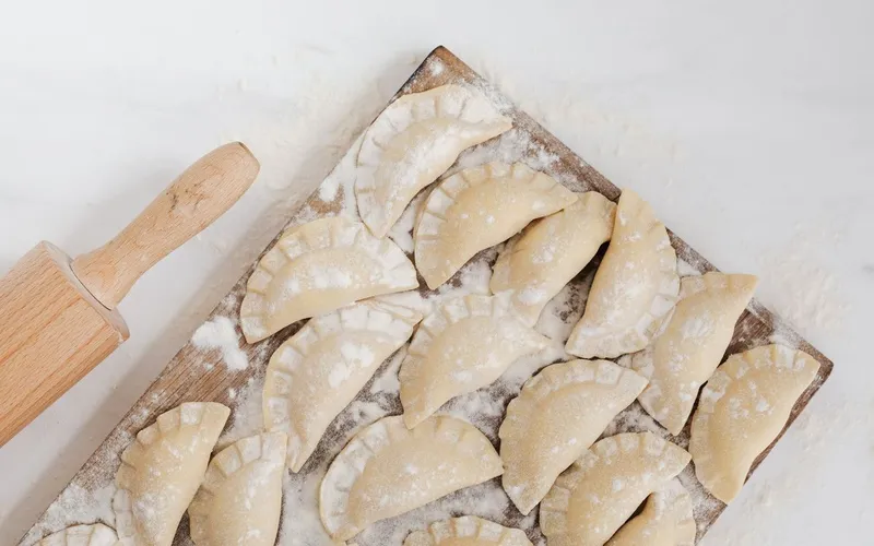 Pierogi crus façonnés à la main, alignés sur une planche farinée, à côté d'un rouleau à pâtisserie en bois