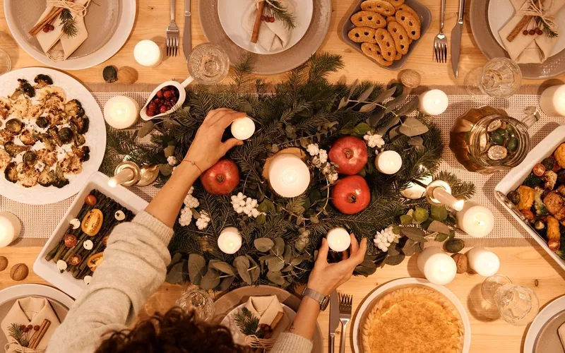 Table de Noël vue du dessus : centre de table en branches de sapin, grenades et bougies, légumes rôtis, tarte, assiettes et serviettes nouées de cannelle