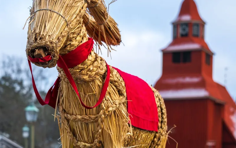 Grand bouc de paille paré d'un ruban et d'une couverture rouges, devant l'église en bois rouge de Seglora au musée de plein air de Skansen, à Stockholm