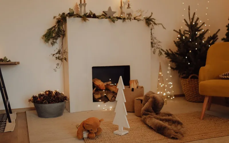 Salon décoré pour Noël : cheminée blanche garnie de bûches, bougies et guirlande de sapin sur le manteau, petit sapin lumineux et ours en peluche sur le tapis